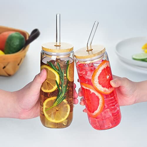 Two hands holding iced drinks in glass jars with fruit slices and straws.